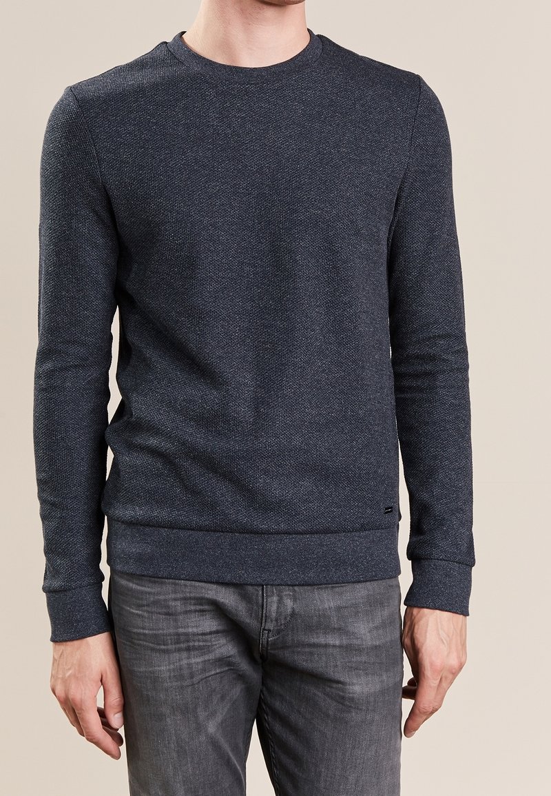 Homme portant un pull gris foncé texturé à manches longues et col rond, et un jean noir délavé, debout devant un fond beige.