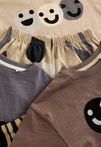 Baumwollkleidungsstücke umfassen ein beiges Oberteil mit Smiley-Gesicht-Drucken und dunklen Akzenten, lila und braune Hemden sowie gemusterte Shorts mit einem elastischen Bund.