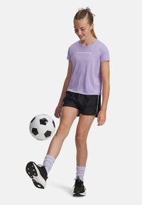 Ung tjej i lila skjorta och svarta shorts som sparkar en svart och vit fotboll mot en enfärgad vit bakgrund.