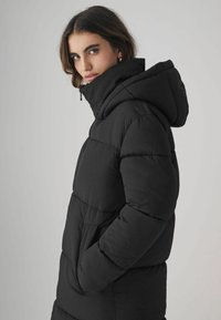 Giacca a vento nera con cappuccio, caratterizzata da un design trapuntato, tasche ampie e una texture sintetica liscia. Elegante e calda.
