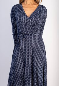 Marinblå omslagsklänning med långa ärmar, vit virvelmönster, V-halsringning och justerbart knytband i midjan; tillverkad av mjukt, stretchigt tyg.