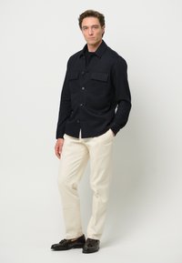 Marineblauwe knoopvoering overhemdjack met twee borstzakken, gecombineerd met romige chino's en zwarte net shoes. Gladde stof met een gestructureerde pasvorm.