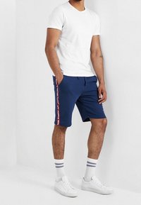 Shorts bleu marine avec un imprimé logo blanc sur le côté, associés à un t-shirt blanc et des baskets blanches, complétés par des chaussettes rayées.