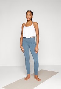 Weißes, gekürztes Tanktop mit dünnen Trägern, kombiniert mit hoch geschnittenen blauen Leggings. Das Model steht barfuß auf einer beigen Yogamatte vor einem schlichten Hintergrund.