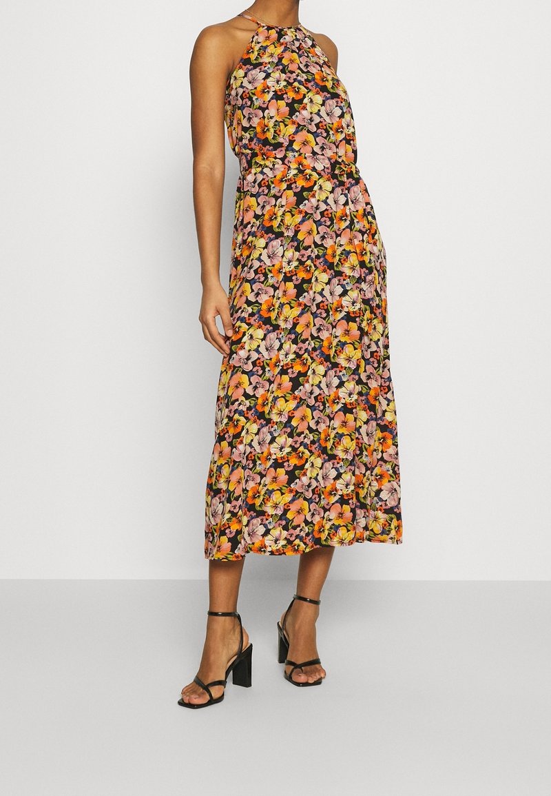 Vestido maxi floral con cuello halter, con un fondo negro y flores rosas y amarillas. Tejido suave y corte holgado. Sandalias de tacón negro.