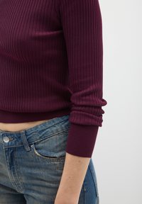 Maglia bordeaux a coste con maniche lunghe e polsini arrotolati, abbinata a jeans in denim blu slavato con tasca frontale e chiusura con bottone.