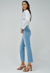 Jeans rasgados de un azul claro con una cintura alta, combinados con sandalias de tacón con tiras marrones y una camisa blanca de botones, vistos desde el lado.