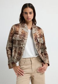 Quiltet brun og beige jakke med en standkrave, der har to frontlommer og et tekstureret mønster, båret over en hvid skjorte.