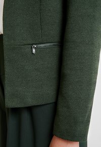 Manche de veste texturée vert foncé avec une petite poche zippée horizontale en argent près du poignet, portée sur un tissu vert foncé.