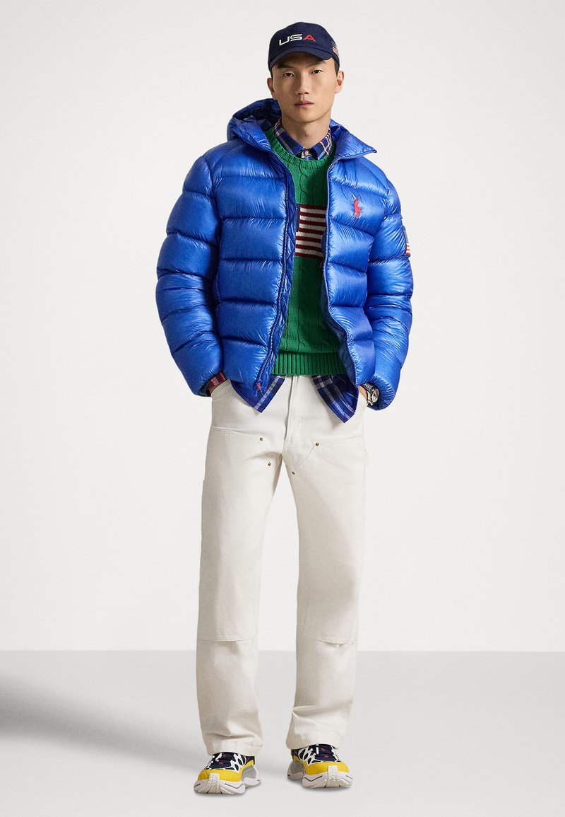 Veste matelassée bleue avec capuche, pull vert à motif rayé, pantalon clair et baskets multicolores. Ensemble décontracté.