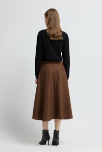 Frau mit glattem, braunem Haar, die ein schwarzes Langarmoberteil, einen braunen Midirock und schwarze, hochhackige Stiefeletten trägt, mit dem Rücken zur Kamera.