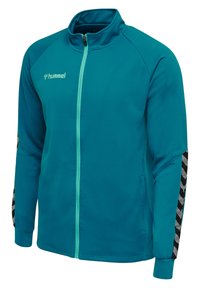 Turkos zip-up jacka gjord av strukturerat tyg, med ett svart och vitt chevronmönster på ärmarna. Logotyp på bröstet, stående krage och figursydda manschetter.