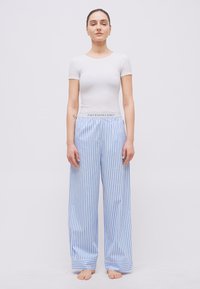 Γυναίκα που στέκεται ξυπόλητη, φορώντας λευκό εφαρμοστό μπλουζάκι και φαρδιά μπλε και λευκή παντελόνα με κάθετες ρίγες και ζώνη με λογότυπο Polo Ralph Lauren.