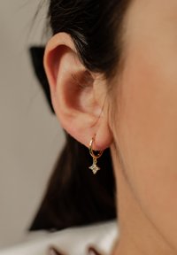 Pendiente de aro dorado con un pequeño charm en forma de estrella que presenta piedras blancas, colgando por debajo del aro, visto en una oreja en primer plano.