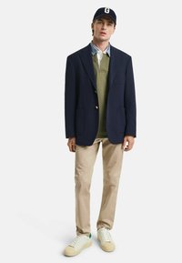 Navy blazer with gold buttons, green collared sweater, white shirt, khaki pants, and white sneakers with green accents. Simple, structured design.