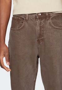 Jean en denim marron avec design classique à cinq poches, coupe droite et texture subtile. Fermeture par bouton métal à la taille.