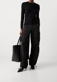 Personne portant un haut noir à manches longues, un pantalon cargo noir ample avec des ourlets à cordon, des talons pointus noirs et tenant un grand sac cabas noir.