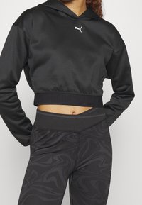 Felpa nera corta con cappuccio e cordoncino, abbinata a leggings neri a vita alta con un motivo scuro a spirale.