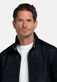 Marineblauwe gewatteerde jas met een staande kraag, voorzien van witte accentstiksels en een rits aan de voorkant. Draag het boven een witte crewneck T-shirt.