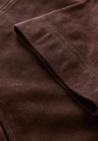 Tissu en daim marron avec une texture lisse, présentant une section pliée et des coutures visibles le long des bords.
