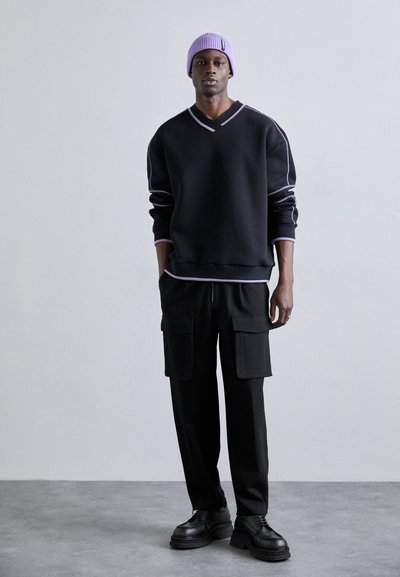 Felpa nera con rifiniture viola, scollatura a V, indossata con pantaloni cargo neri dotati di due ampie tasche. A completare il look ci sono scarpe nere e un berretto viola.