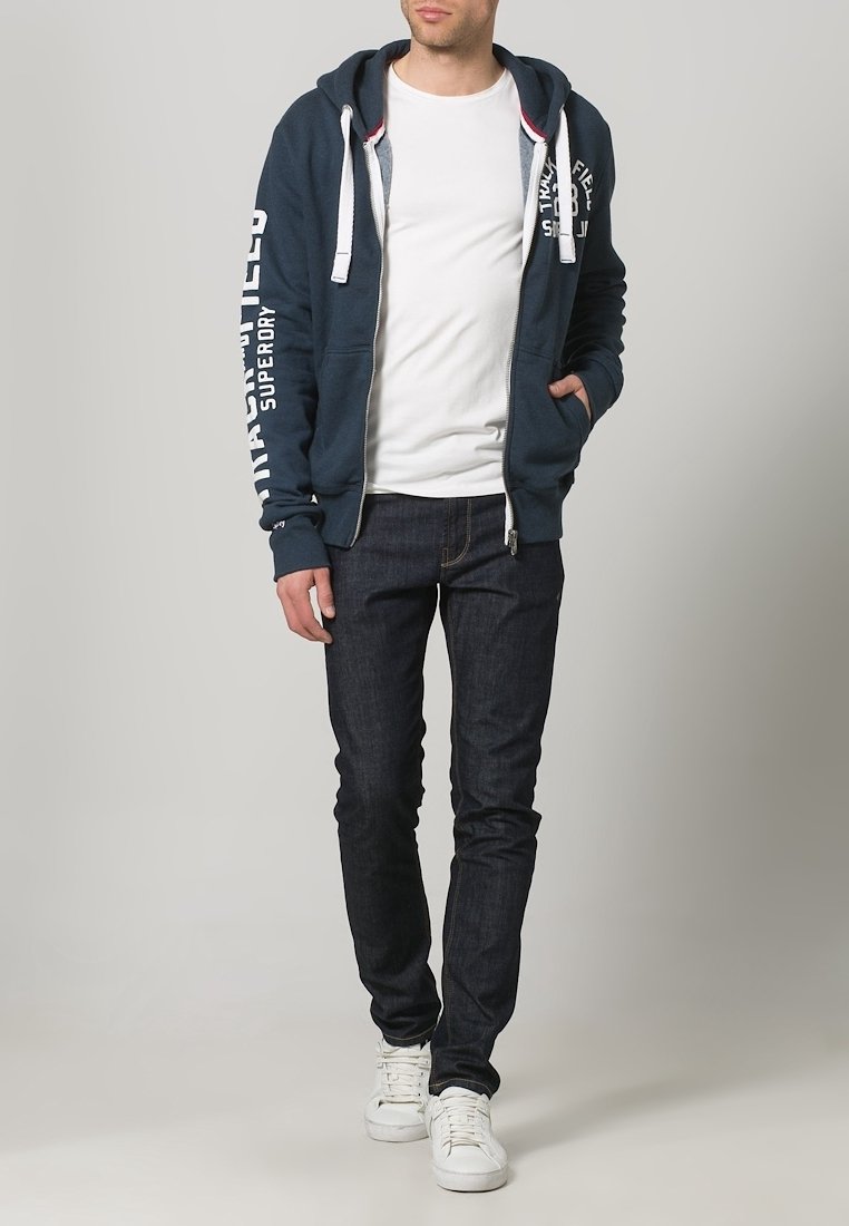 Hombre vistiendo una sudadera con cremallera en azul marino con texto blanco, camiseta blanca, jeans oscuros y zapatillas blancas, de pie con la mano en el bolsillo.