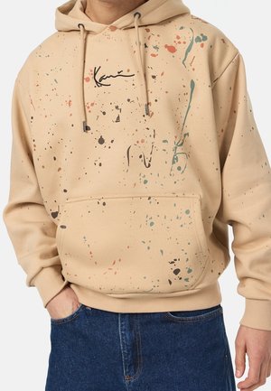 Beigefarbener Kapuzenpullover mit buntem Farbspritzmuster, Vorder Tasche und schwarzem kursivem Text, getragen zu blauen Jeans.
