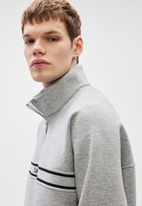 Γκρι φούτερ με φερμουάρ τεταρτημόριου και ψηλό γιακά, με λευκή και μαύρη ριγέ λεπτομέρεια και λογότυπο της μάρκας στο στήθος. Μαλακό, υφασμένο ύφασμα.