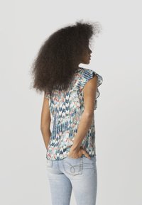 Farebná blúzka s kombinovanými vzormi a volánovými rukávmi. Nosí sa s svetlomodrými džínsami, ktoré zvýrazňujú textúrovanú tkaninu.