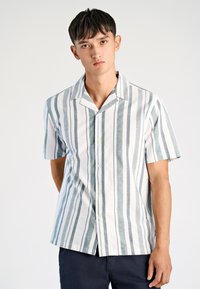 Jonge man met kort, donker haar draagt een wit overhemd met korte mouwen met verticale groene strepen en donkere broek, staand tegen een effen achtergrond.