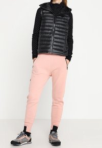 Mustat paksut liivimuotoiset ilmanhihoja oleva takki mustan kuvioidun pitkähihaisen paidan päällä, yhdistettynä vaaleanpunaisiin verkkareihin ja harmaisiin urheilukengiin.