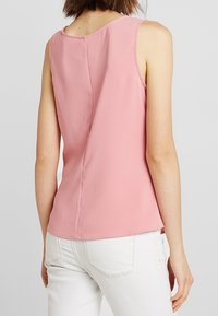 Top sin mangas rosa con un escote redondeado, textura suave y una costura central en la parte trasera, combinado con pantalones blancos.