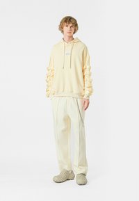 Sudadera amarilla con acentos prominentes en forma de cintas en las mangas, combinada con pantalones claros amarillos ajustados y zapatos beige texturizados.