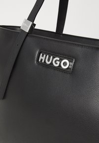 Czarna skórzana torba typu tote o usztywnionym kształcie. Posiada srebrną naszywkę z logo "HUGO" oraz eleganckie metalowe okucia. Gładkie, teksturowane wykończenie.