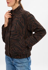 Woman wearing a brown and black tiger print jacket with a high collar, hand in pocket, and blue jeans against a plain white background.