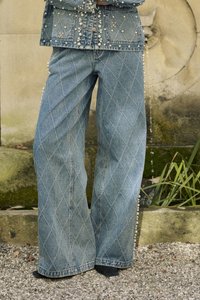 Džínsy s diamantovým vzorom stehu, spárované s denimovou bundou zdobenou perlami, stojace na štrku pri kamennom fontáne.