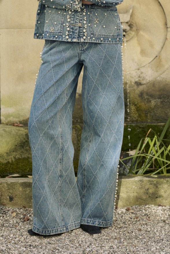 Džínsy s diamantovým vzorom stehu, spárované s denimovou bundou zdobenou perlami, stojace na štrku pri kamennom fontáne.