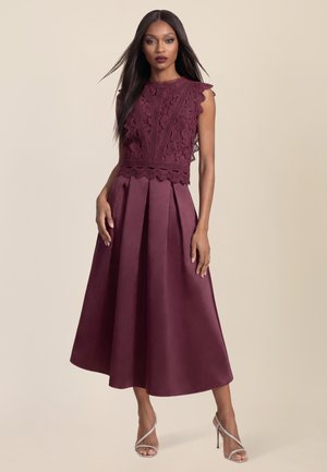 Frau trägt ärmelloses bordeauxrotes Spitzenoberteil und passenden plissierten Midirock mit silbernen Riemchenabsätzen, steht vor einfachem Hintergrund.