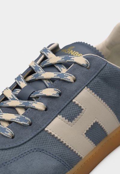 Κοντινό πλάνο αθλητικού παπουτσιού σε μπλε και μπεζ χρώμα με υφή υφάσματος, κορδόνια με σχέδιο, μπεζ λογότυπο "H" στο πλάι και καφέ ελαστική σόλα.