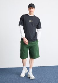 Mustat Nike-t-paidat, jossa on kultainen logo, valkoinen pitkähihainen paita alla, vihreät shortsit ja valkoiset lenkkarit. Minimalistinen muotoilu, rento istuvuus.