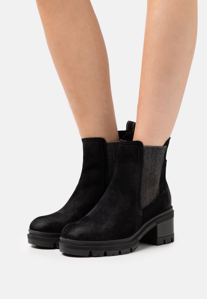 Bottes cheville en daim noir avec talons épais et panneaux latéraux texturés nervurés portées sur des jambes nues contre un fond blanc.