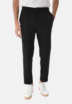 Uomo con pantaloni neri attillati con coulisse, risvoltini, sneakers bianche e una camicia bianca a maniche corte, mani in tasca.