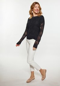Schwarzer gestrickter Pullover mit geometrischem Spitzenmuster, weiße, enganliegende Hose und beigefarbene Mokassins mit goldenen Akzenten. Das Modell posiert leicht gedreht.
