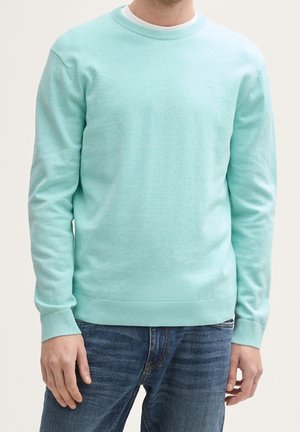 Homme portant un pull à manches longues bleu clair sur un sous-vêtement blanc avec un jean en denim bleu, debout devant un fond uni.