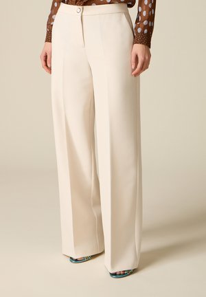 Vrouw draagt een high-waist, wijde crème broek en open tenen gestreepte hakken, gecombineerd met een bruine blouse met lange mouwen en polkadots.