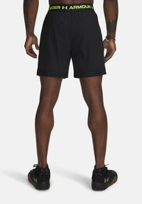 Schwarze Sportshorts mit einem markierten neon-gelben Bund, flach geschnitten und aus einem leichten, glatten Material. Getragen mit schwarzen Turnschuhen.