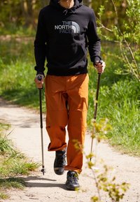 Persona che cammina su un sentiero di terra con bastoni da trekking, indossando pantaloni arancioni, felpa nera della North Face e scarpe da trekking nere.