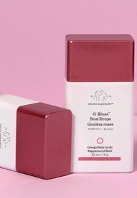 Square-shaped bottle with a matte white base and a shiny rose gold lid. Label reads "O-Bloos™ Rosi Drops," 30 ml. Background is light pink.