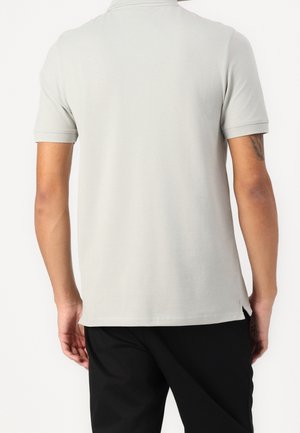 Personne portant une chemise à manches courtes unie gris clair et un pantalon noir, vue de dos sur fond blanc.