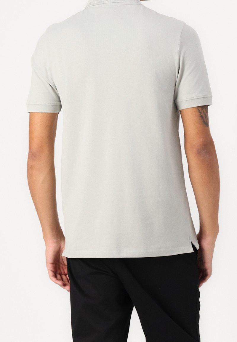 Personne portant une chemise à manches courtes unie gris clair et un pantalon noir, vue de dos sur fond blanc.